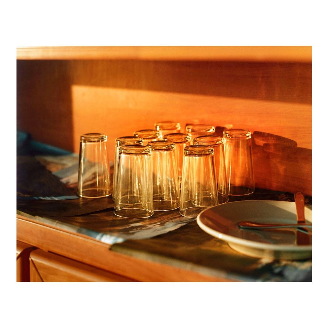 Still life photograph of a collection of empty drinking glasses and a plate with a spoon on a wooden surface, bathed in warm lighting.