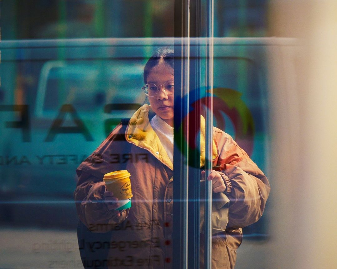 Woman in a jacket stands near a window holding a beverage; an emergency vehicle with safety text is visible in the window's reflection.