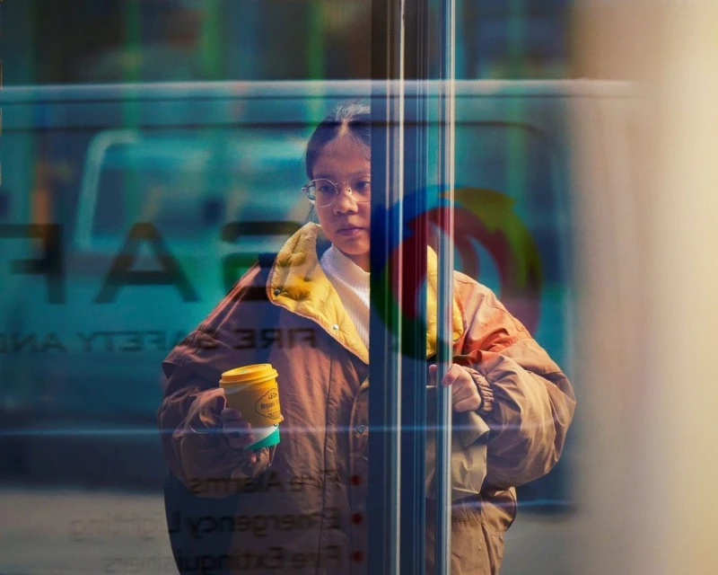 Woman in a jacket stands near a window holding a beverage; an emergency vehicle with safety text is visible in the window's reflection.
