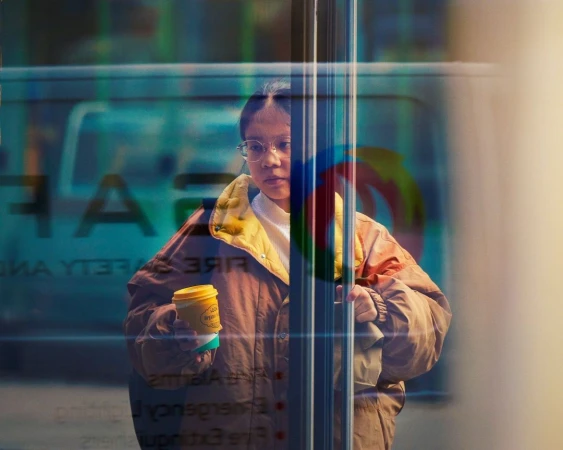 Woman in a jacket stands near a window holding a beverage; an emergency vehicle with safety text is visible in the window's reflection.