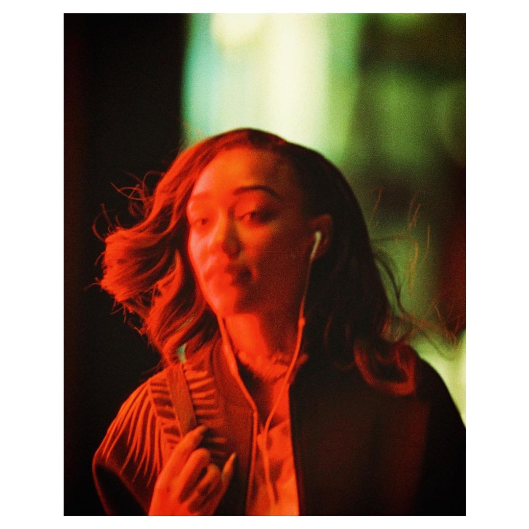 A young woman is seen in a close-up outdoor portrait, set against an abstract background, with dominant red and green hues under dim lighting.