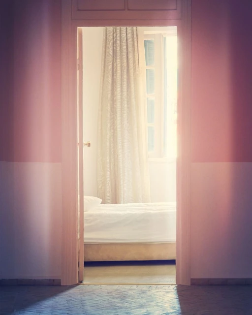 Bedroom interior seen through open doorway; white bed linens, sheer curtains, and light-pink walls create a soft, bright, clean aesthetic.
