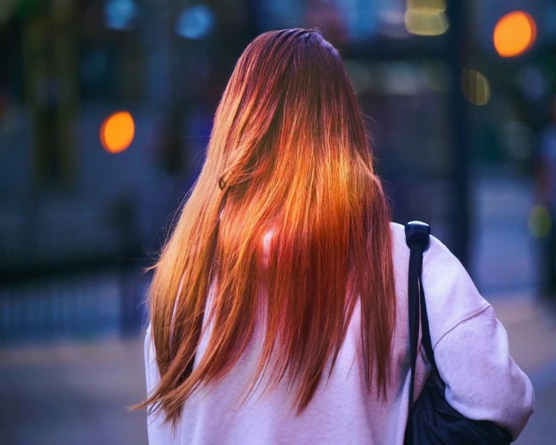 Back view of a red-haired woman in a sweatshirt walking in an urban setting with blurred lights in the background during twilight hours.