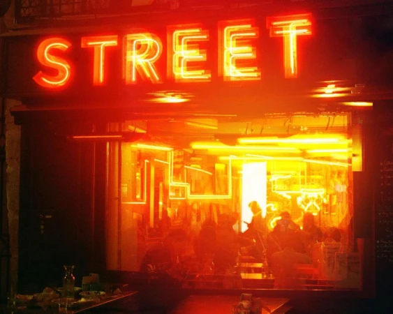 Neon sign reading 'STREET' illuminates a dimly lit restaurant interior with patrons, tables, and a bar visible in the background.