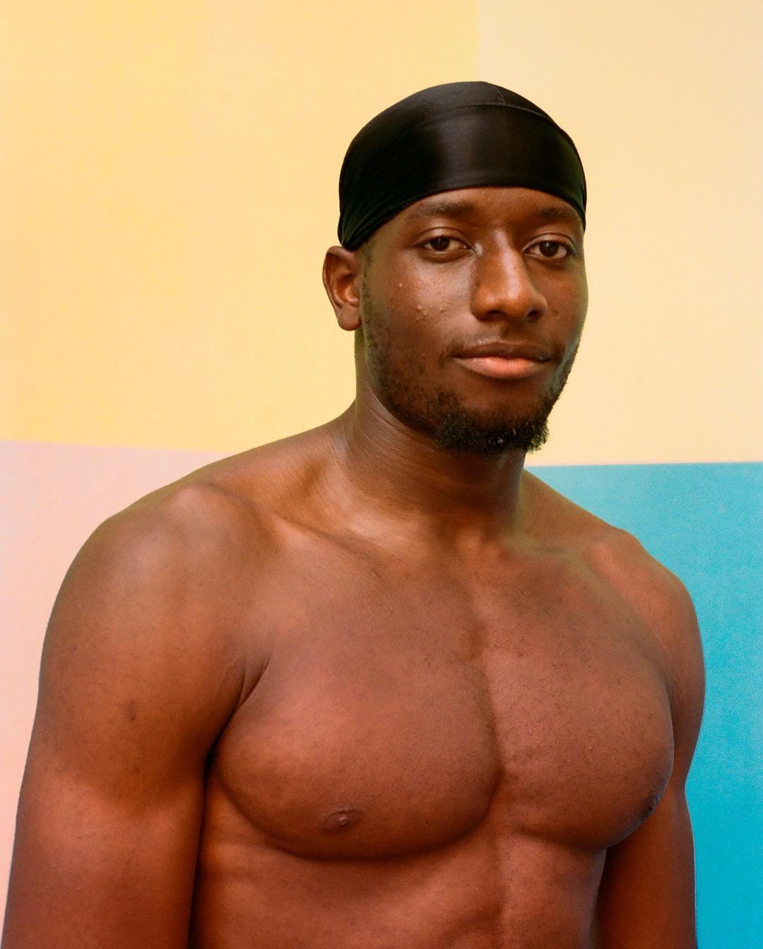 Portrait of a shirtless African-American male with a black durag against a colorful backdrop, showcasing fitness and personal style.