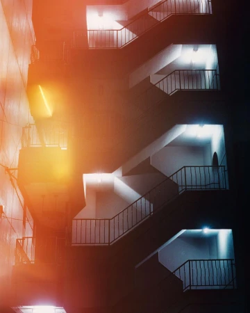 Architectural nighttime exterior of a modern building featuring an emergency staircase with iron railings and electric lighting.