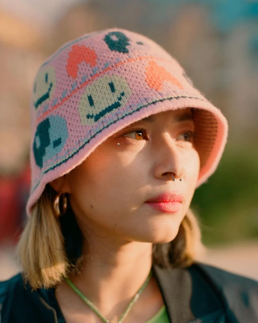 Young woman wearing a pink patterned bucket hat looks at the camera with soft sunlight illuminating her face in an outdoor setting.