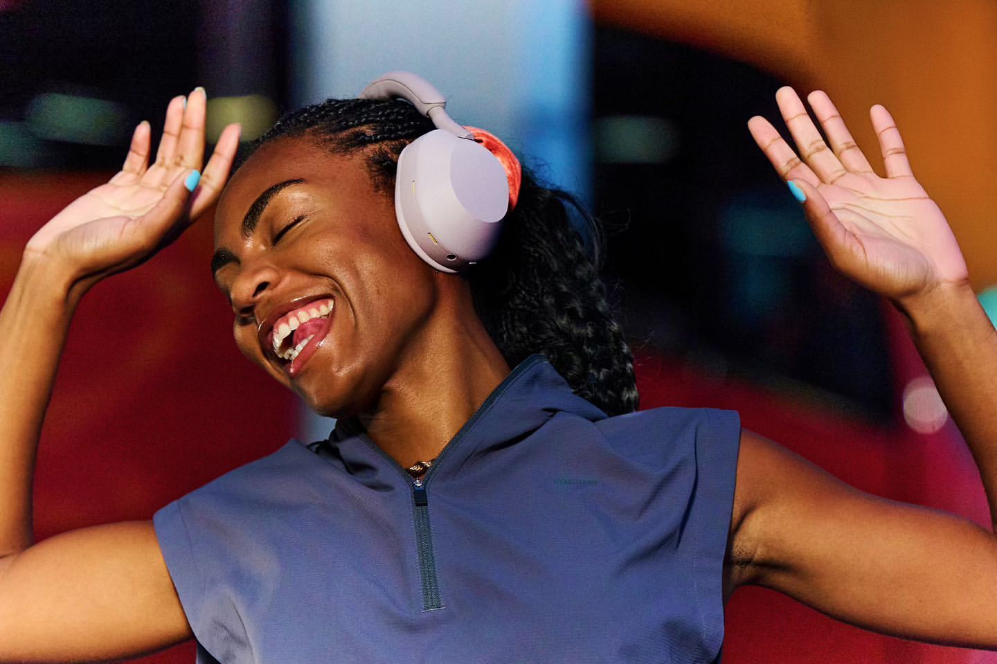 Woman with wireless headphones laughs with her eyes closed and her arms raised in an indoor environment with bright colors.