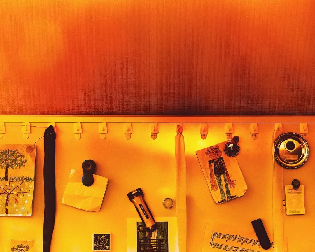 Yellow bulletin board with children's drawings, notes, and small objects attached. Top part is a textured orange wall.