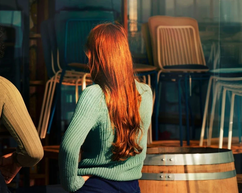 Back view of a person with long red hair in a light-green sweater, standing near stacked chairs and a wooden barrel indoors.
