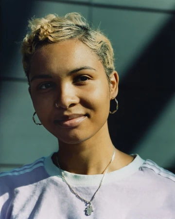 Portrait of a young woman with short dyed blonde and dark hair wearing hoop earrings, a silver necklace, and a pale top.