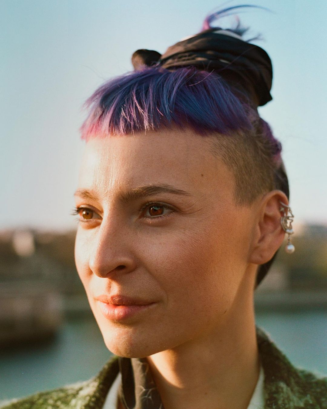 Close-up portrait of a woman with shaved sides, vibrant purple hair, wearing hoop earring against a bright outdoor skyline background.