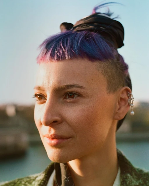 Close-up portrait of a woman with shaved sides, vibrant purple hair, wearing hoop earring against a bright outdoor skyline background.