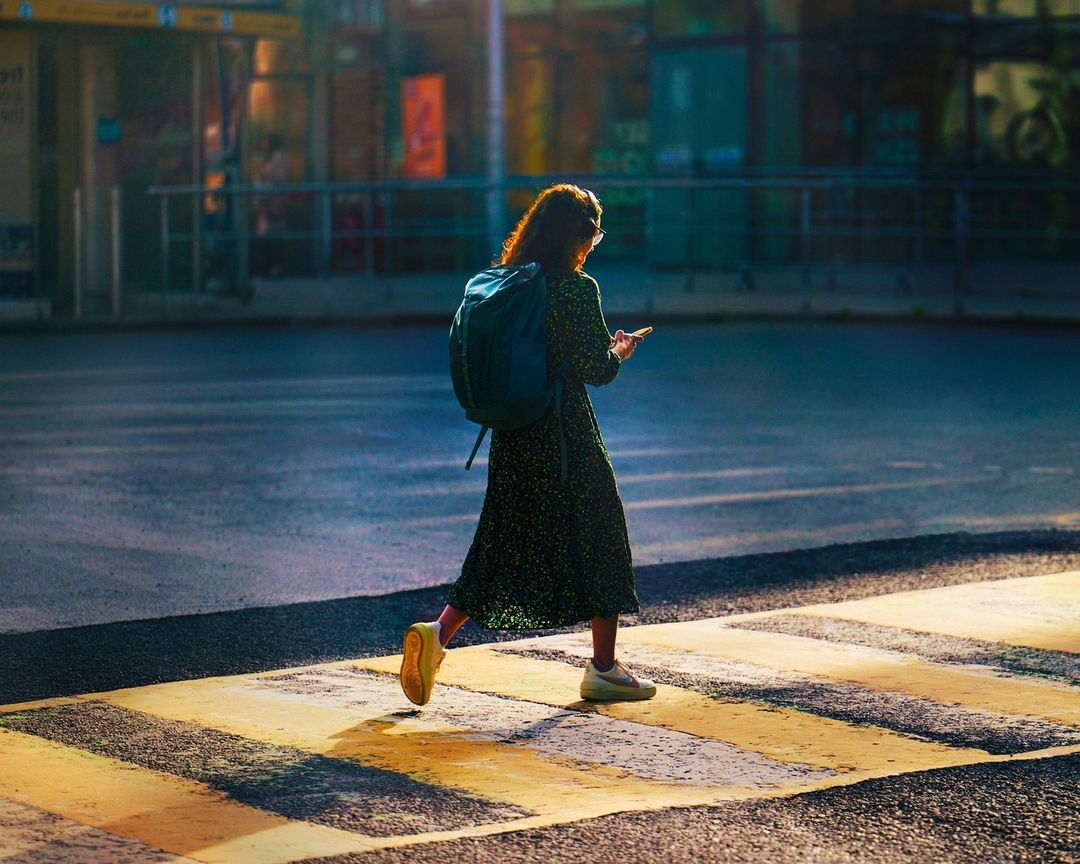 A woman in a patterned dress and backpack is walking across a crosswalk while looking at her phone on a sunny street.