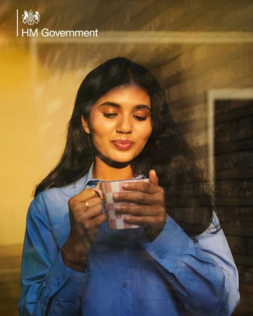 Woman in blue shirt holds checkered mug indoors, bathed in golden sunlight near a window, creating a warm and inviting atmosphere.