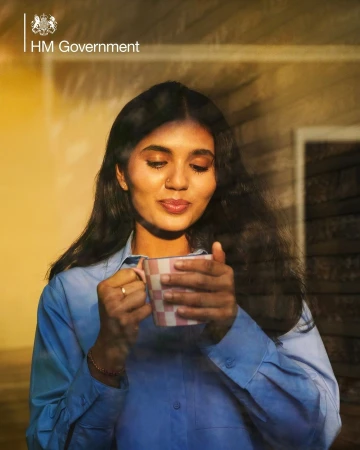 Woman in blue shirt holds checkered mug indoors, bathed in golden sunlight near a window, creating a warm and inviting atmosphere.
