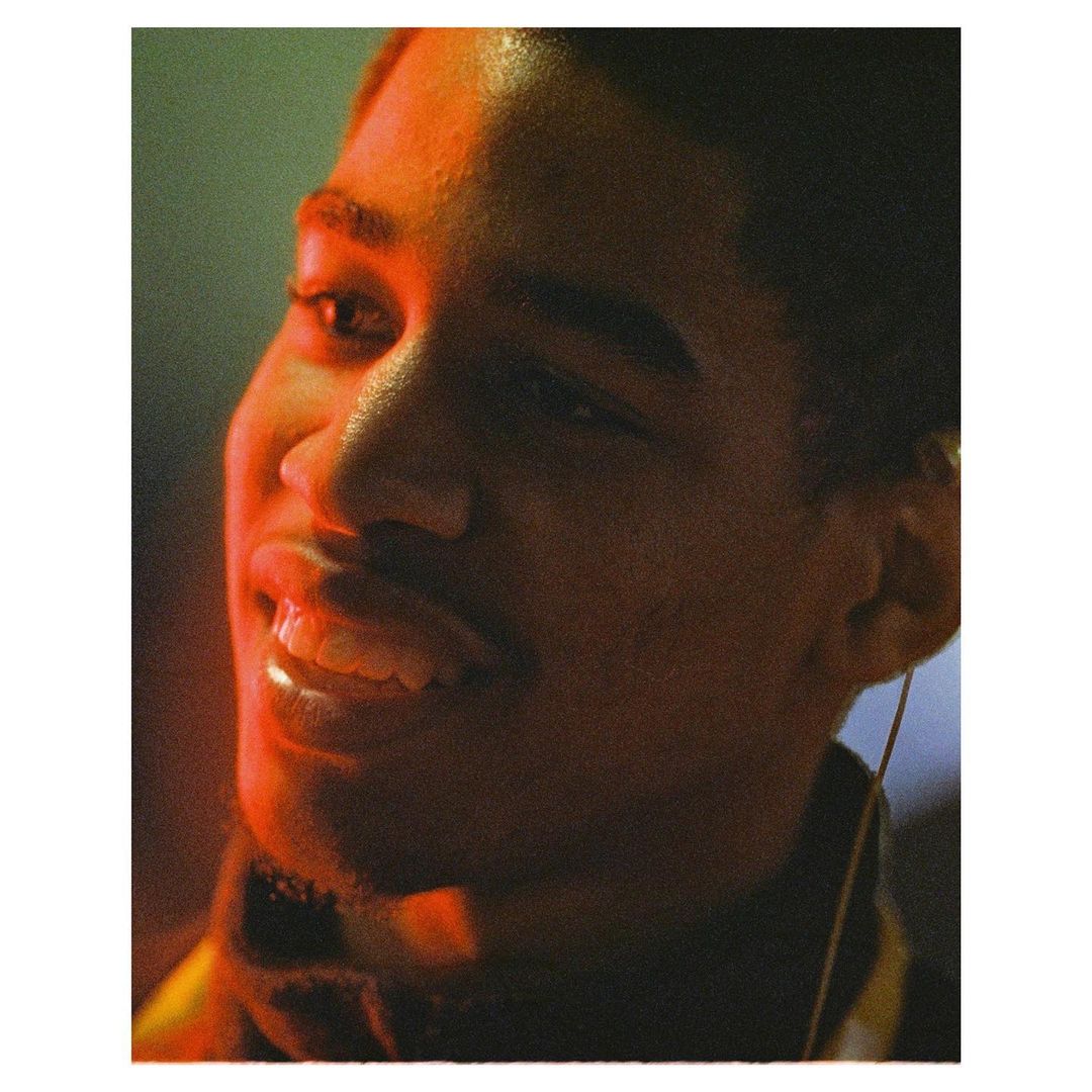 Close-up portrait of a smiling young adult male with short hair, bathed in warm orange light against a dark, out-of-focus green background.