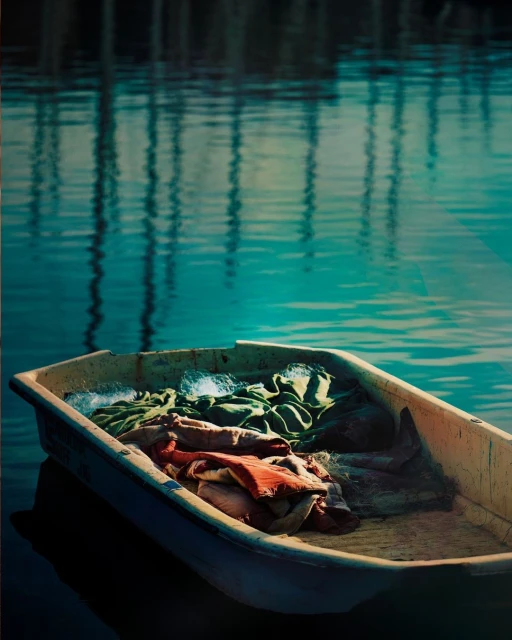 Old rowboat filled with cloth rests on calm turquoise water, reflecting wooden pilings in the background on a sunny day.