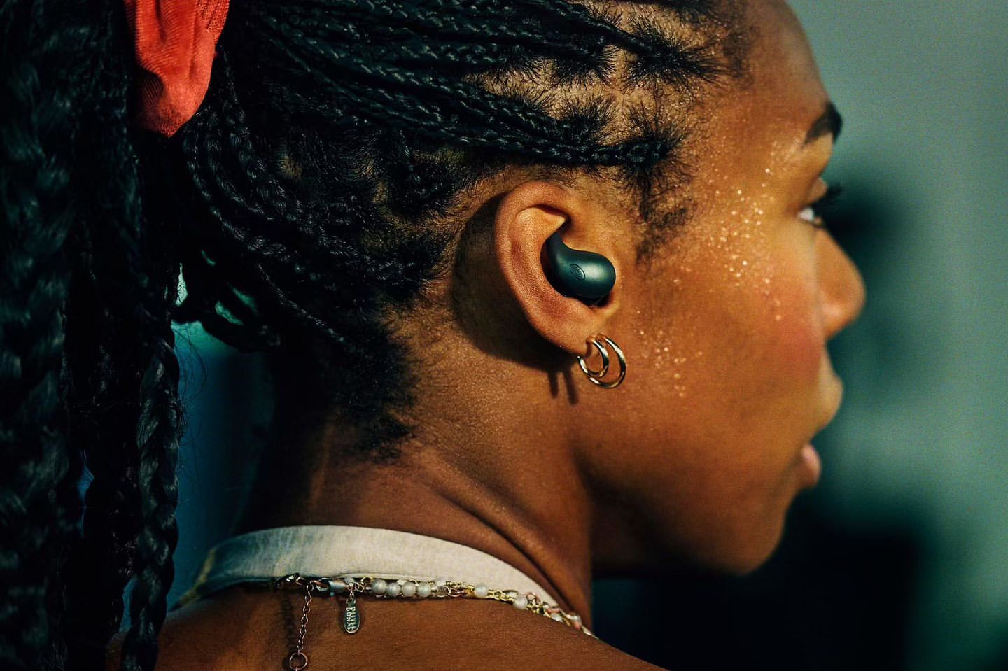 Close-up of a woman with dark braids and an earbud in her ear, wearing gold hoop earrings. Beauty shot highlighting jewelry and technology.