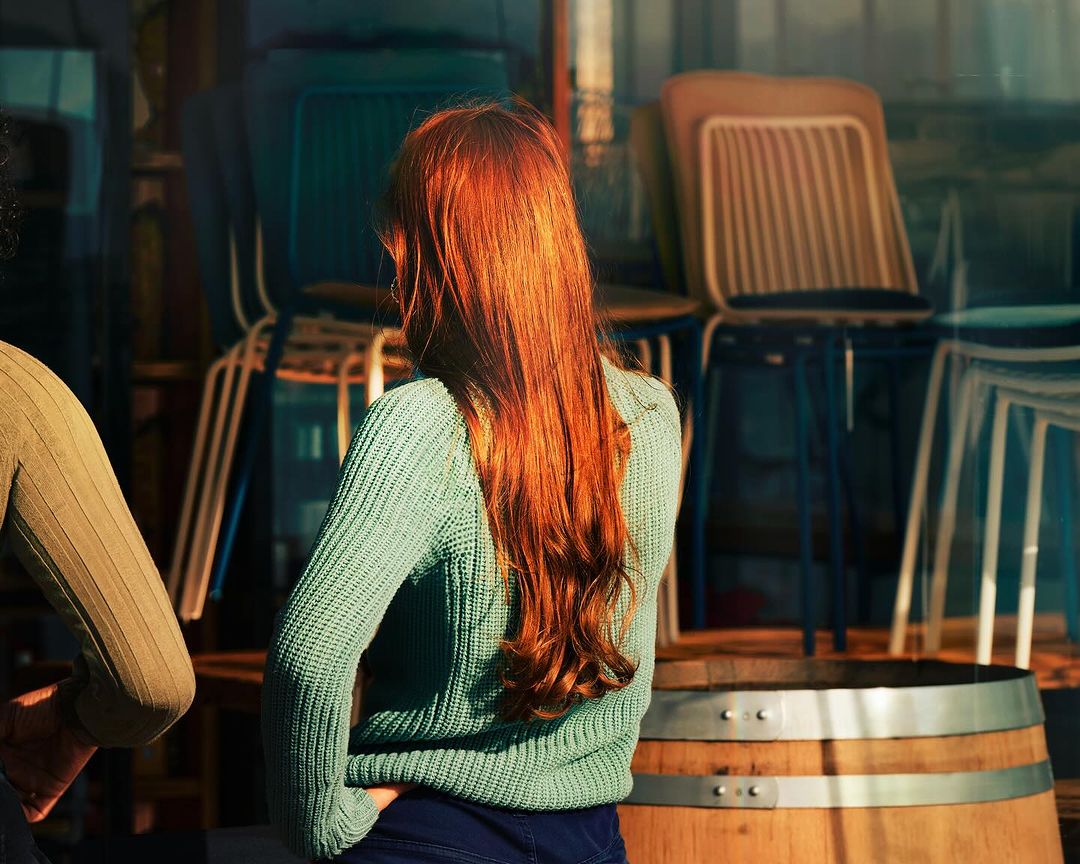 Back view of a woman with long red hair wearing a green sweater, standing near a wooden barrel with chairs stacked in the background.