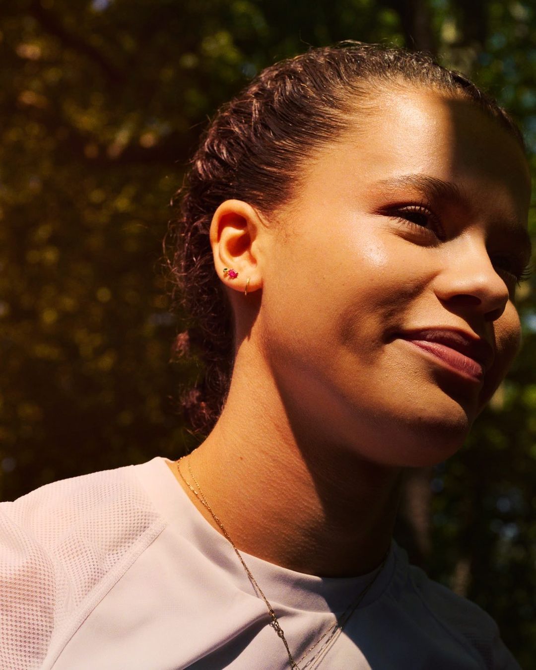 Close-up of a young woman with braided hair and a white athletic shirt outdoors in a sunlit forest, showcasing healthy skin and earrings.
