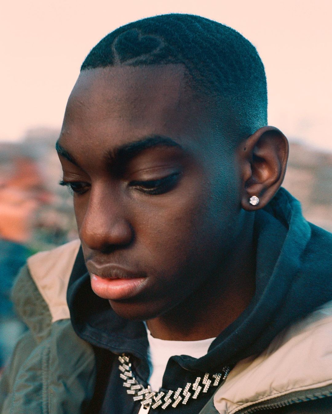 Close-up portrait of a young Black man with a shaved heart design in his hair, wearing a silver chain and hoodie, by Ben Stockley.