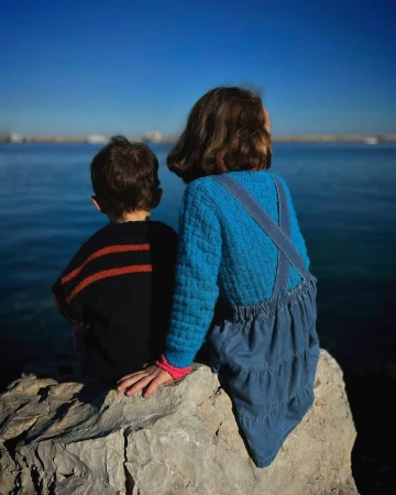 Two children sitting on a rock by the ocean, seen from behind under a clear blue sky