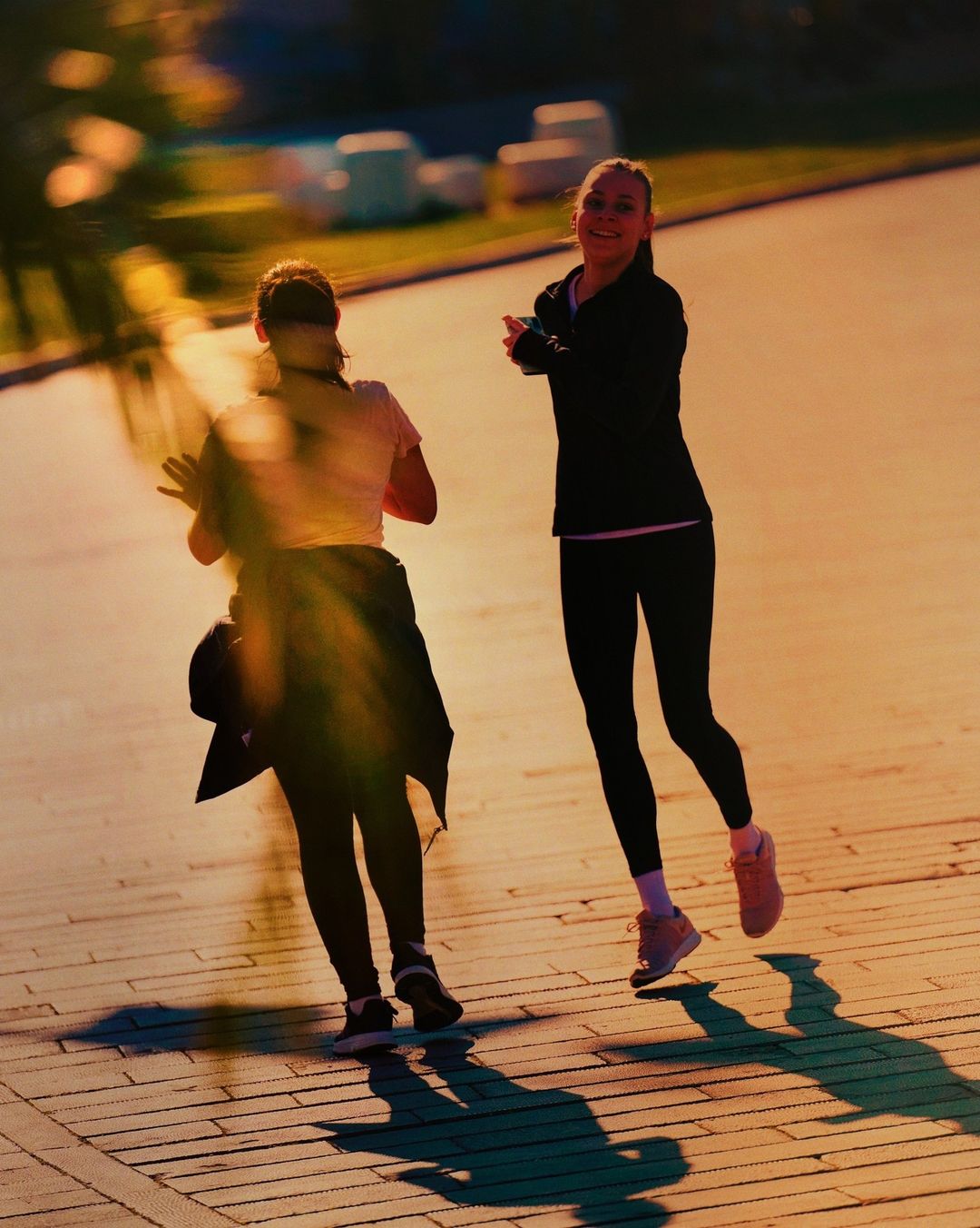 Two women in athletic wear walking on a brick path with shadows, backlit by golden light, smiles, and motion blur.