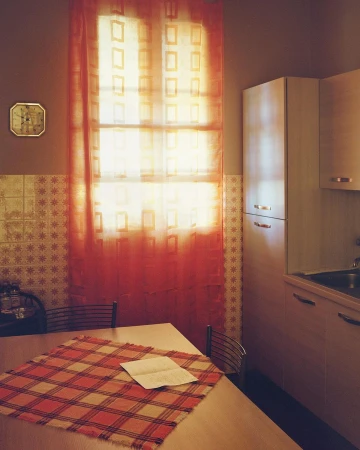 Indoor scene with a table covered by a red-and-beige tablecloth, a window with an orange curtain, and kitchen cabinets.