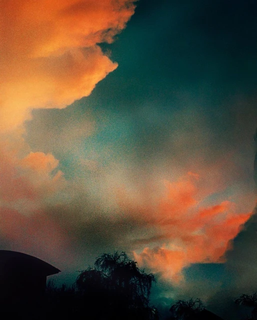 Dramatic skyscape featuring vivid orange clouds against a deep teal sky, with silhouetted rooftops and trees visible in the foreground.