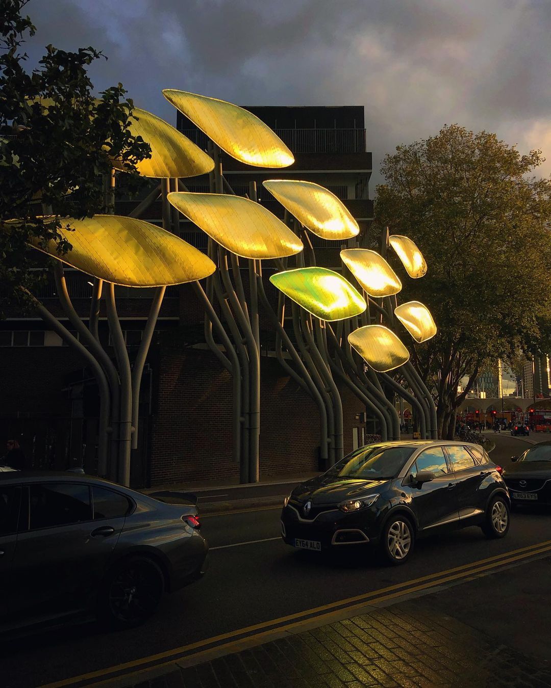 Street scene at dusk featuring a public art installation with leaf-like shapes, silhouetted trees, and cars on the roadway.