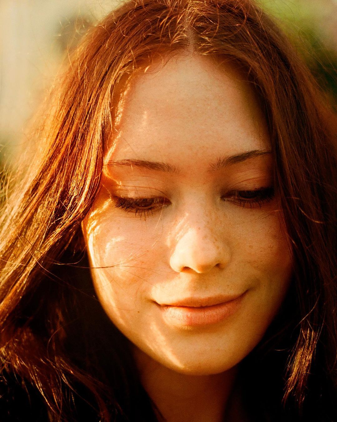 Close-up portrait of a young adult woman with red hair and freckles looking down with a slight smile in soft, warm lighting.
