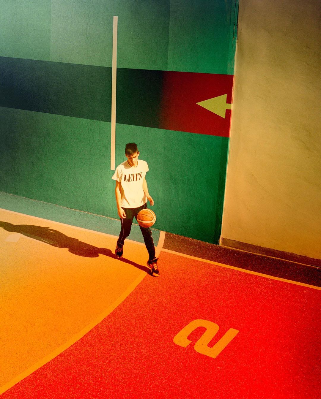 Teenage boy bounces basketball on indoor court featuring green wall with red arrow, floor with orange and red sections and large '2' marking.
