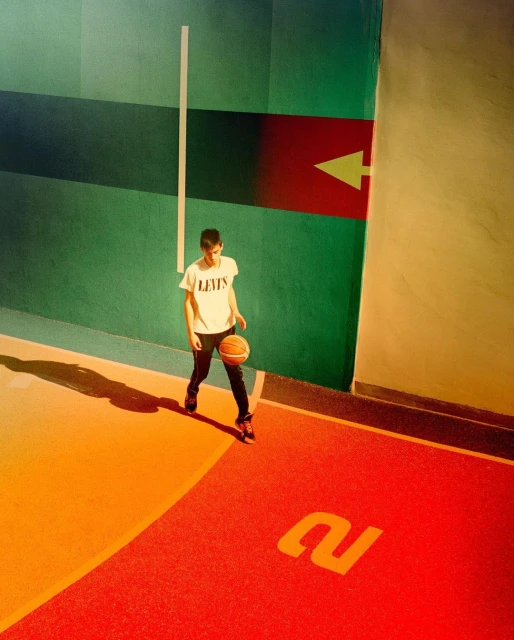 Teenage boy bounces basketball on indoor court featuring green wall with red arrow, floor with orange and red sections and large '2' marking.