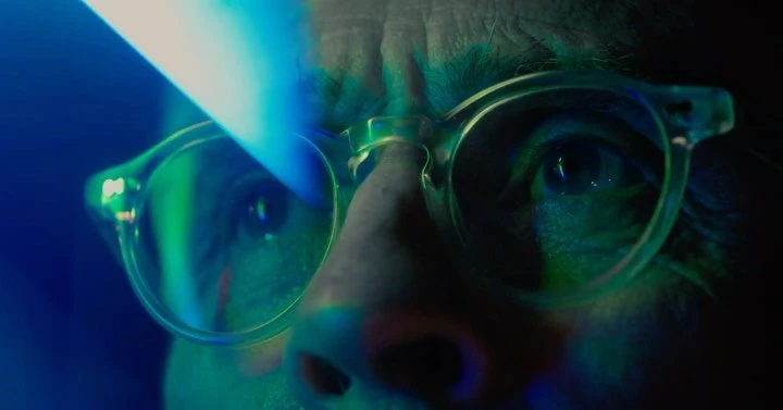 Close-up of an adult wearing eyeglasses with blue and green lighting reflecting on the lens. The image is shot with a shallow depth of field.