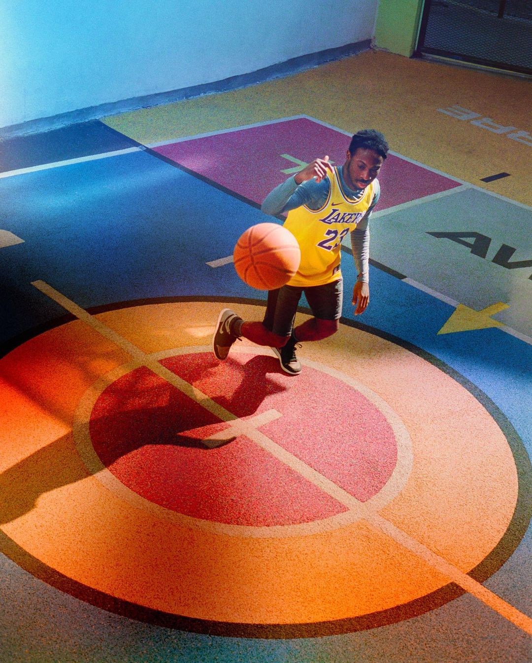 Man in Los Angeles Lakers jersey dribbles basketball on a colourful indoor court with circles and geometric patterns, shot from high angle.