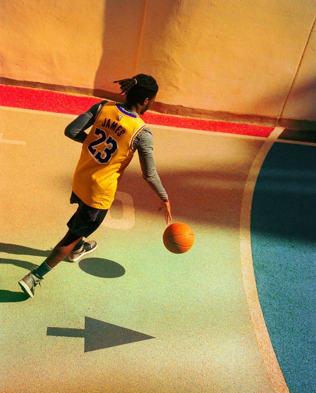 An athlete in a basketball jersey dribbles a basketball on a colorful court with a painted arrow, illuminated by bright, warm lighting.