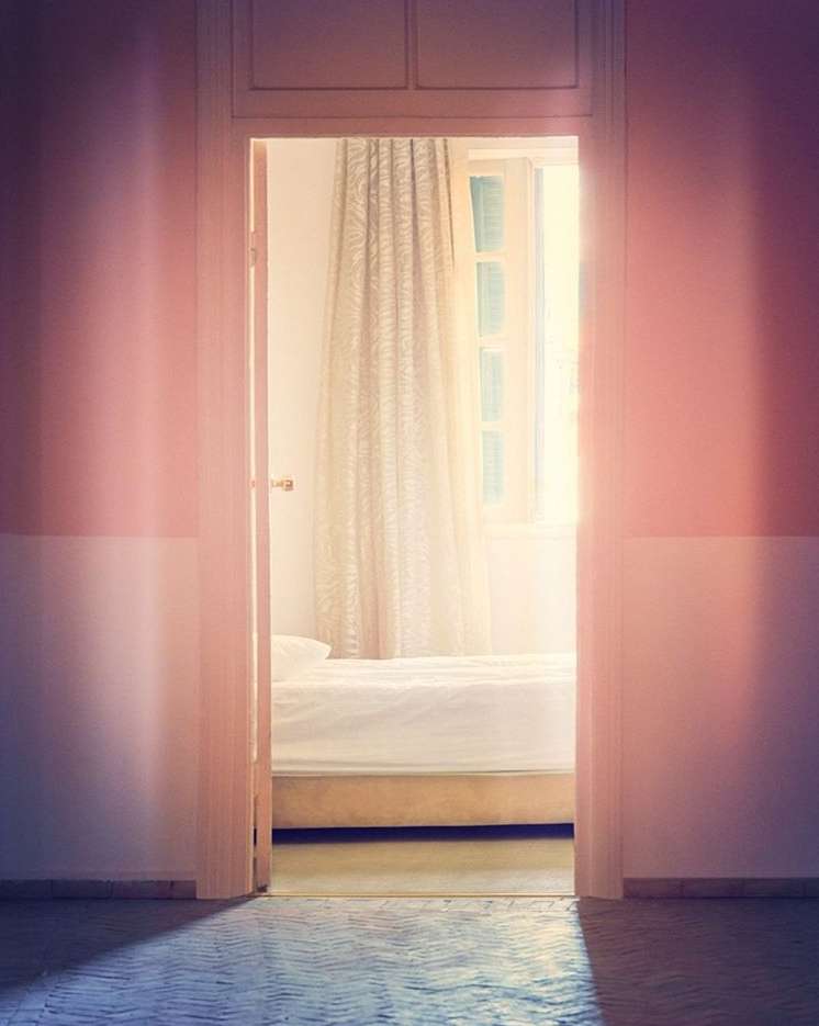 A bedroom is viewed through a doorway; soft lighting and pastel tones create a calm, minimalist aesthetic with a white bed and window.