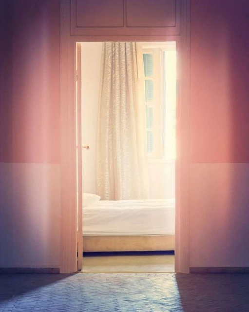 A bedroom is viewed through a doorway; soft lighting and pastel tones create a calm, minimalist aesthetic with a white bed and window.