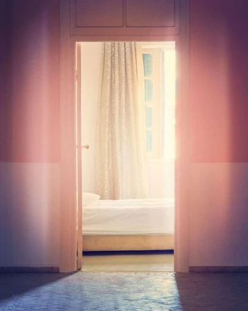 A bedroom is viewed through a doorway; soft lighting and pastel tones create a calm, minimalist aesthetic with a white bed and window.