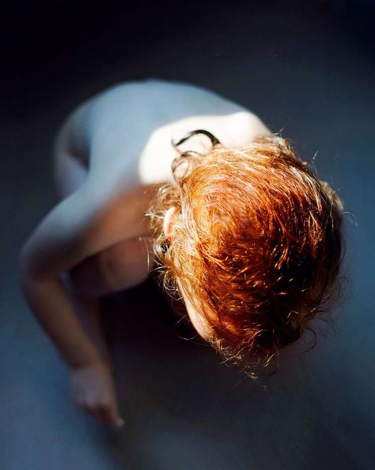 Top-down view of a fair-skinned adult woman with curly red hair, kneeling in diffused light against a cool-toned dark background.