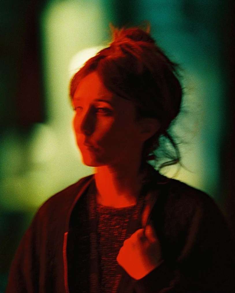Portrait of a woman with her hair in a bun, lit with red and green light in an indoor setting, wearing a dark jacket.