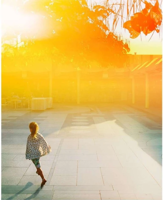 Back view of a child in patterned poncho walking across stone courtyard on a bright, sunny day with intense yellow sunlight and shadows.