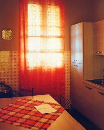 Interior shot of a kitchen featuring a table with a plaid tablecloth, a window with an orange patterned curtain, and kitchen cabinets.