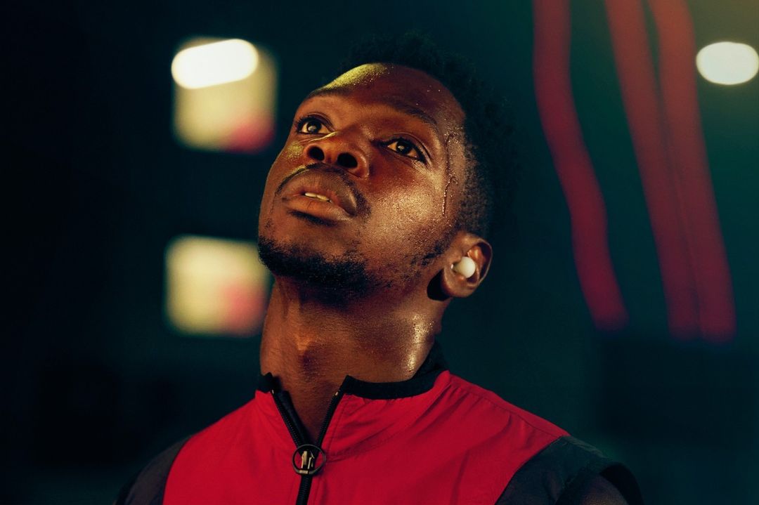 Close-up of a Black man with short hair, wearing a red jacket and earbuds, looking up in an indoor, dark, blurry environment.