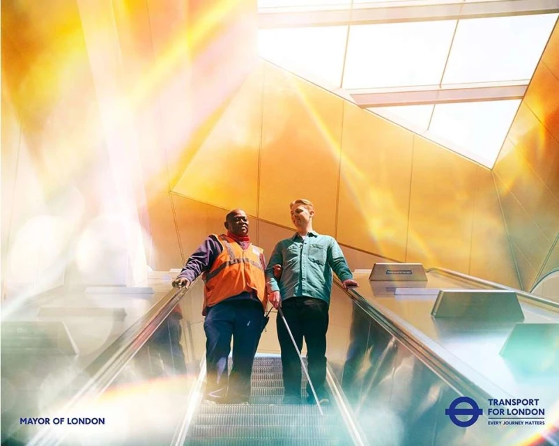 Two men on an escalator, one appearing to assist the other with a white cane, in an indoor public space with bright ambient lighting.