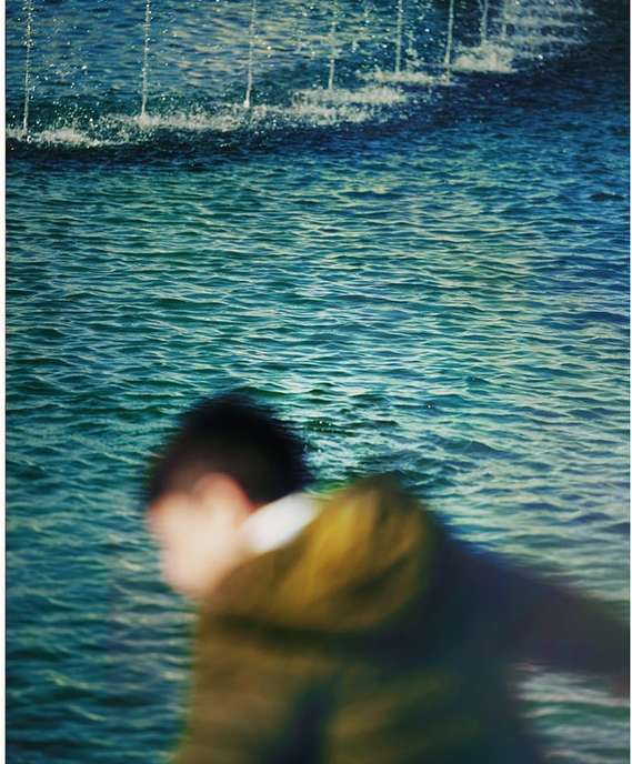 Blurred motion of a person walking near a rippling body of water with a fountain spraying water upward in an outdoor environment.