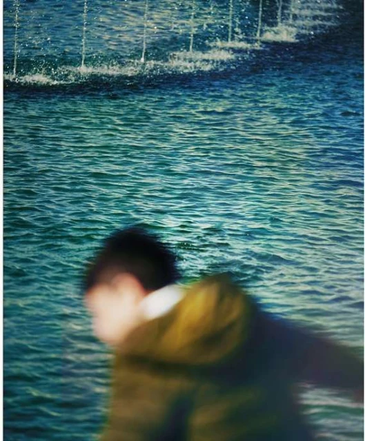 Blurred motion of a person walking near a rippling body of water with a fountain spraying water upward in an outdoor environment.