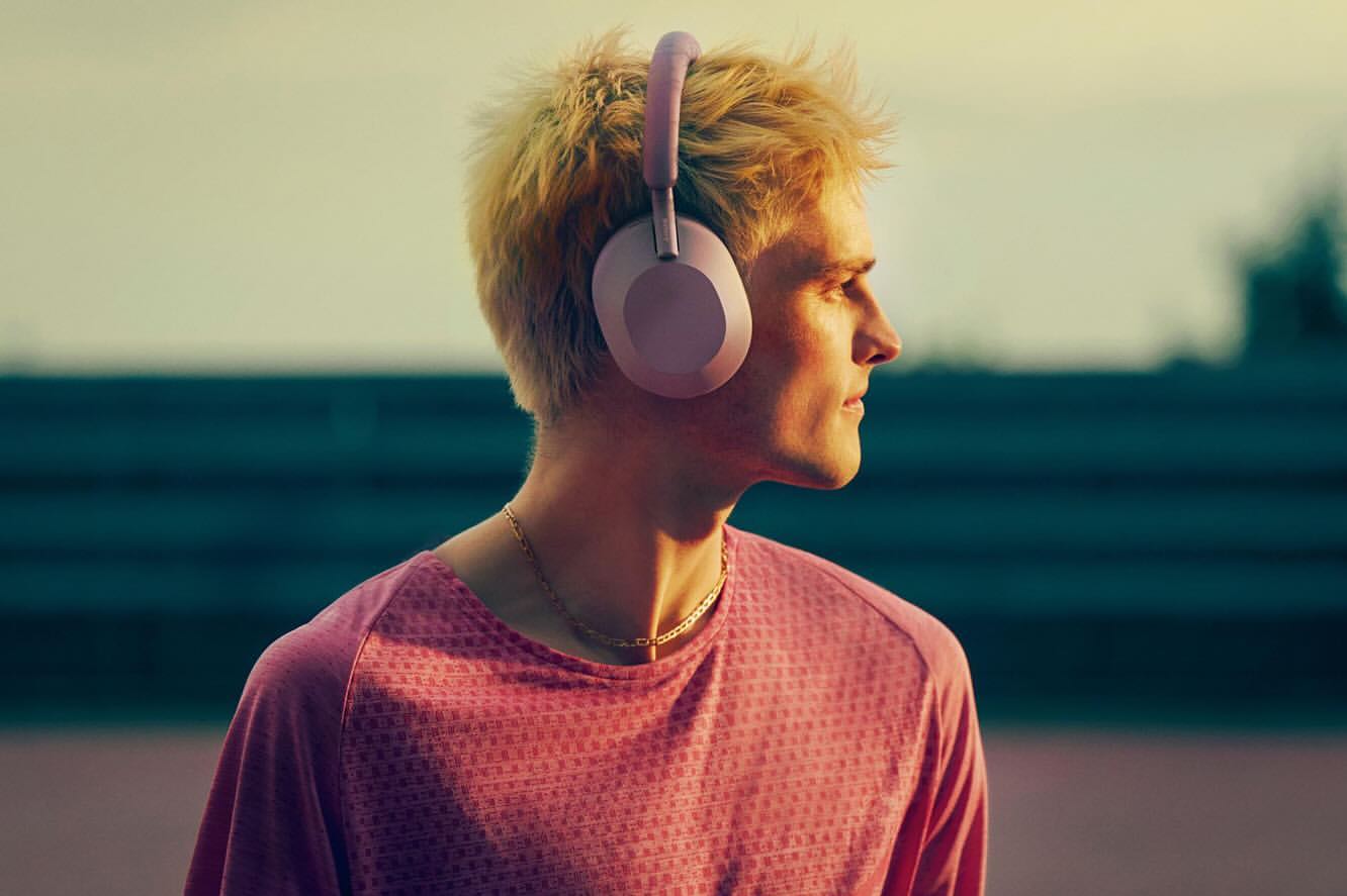 Profile shot of a fair-skinned male wearing mauve headphones and a patterned pink shirt with a neutral background outdoors.
