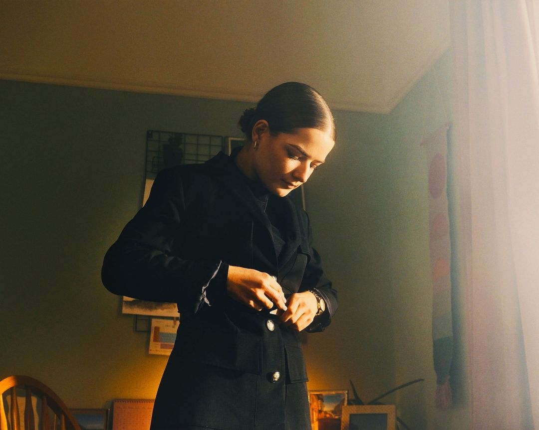 Woman in a black blazer buttoning her jacket in an indoor setting with muted light and pale green walls and framed art.